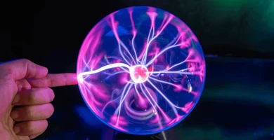 A hand touching a plasma globe, with glowing pink and purple filaments reaching from the center to the glass.
